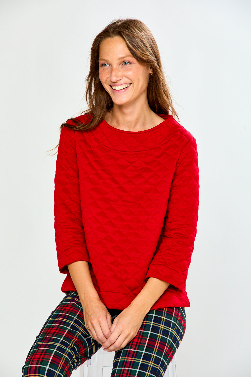 A woman modeling a red long-sleeve quilted knit top with a boat neckline, paired with green and red plaid pants, against a light gray studio background.