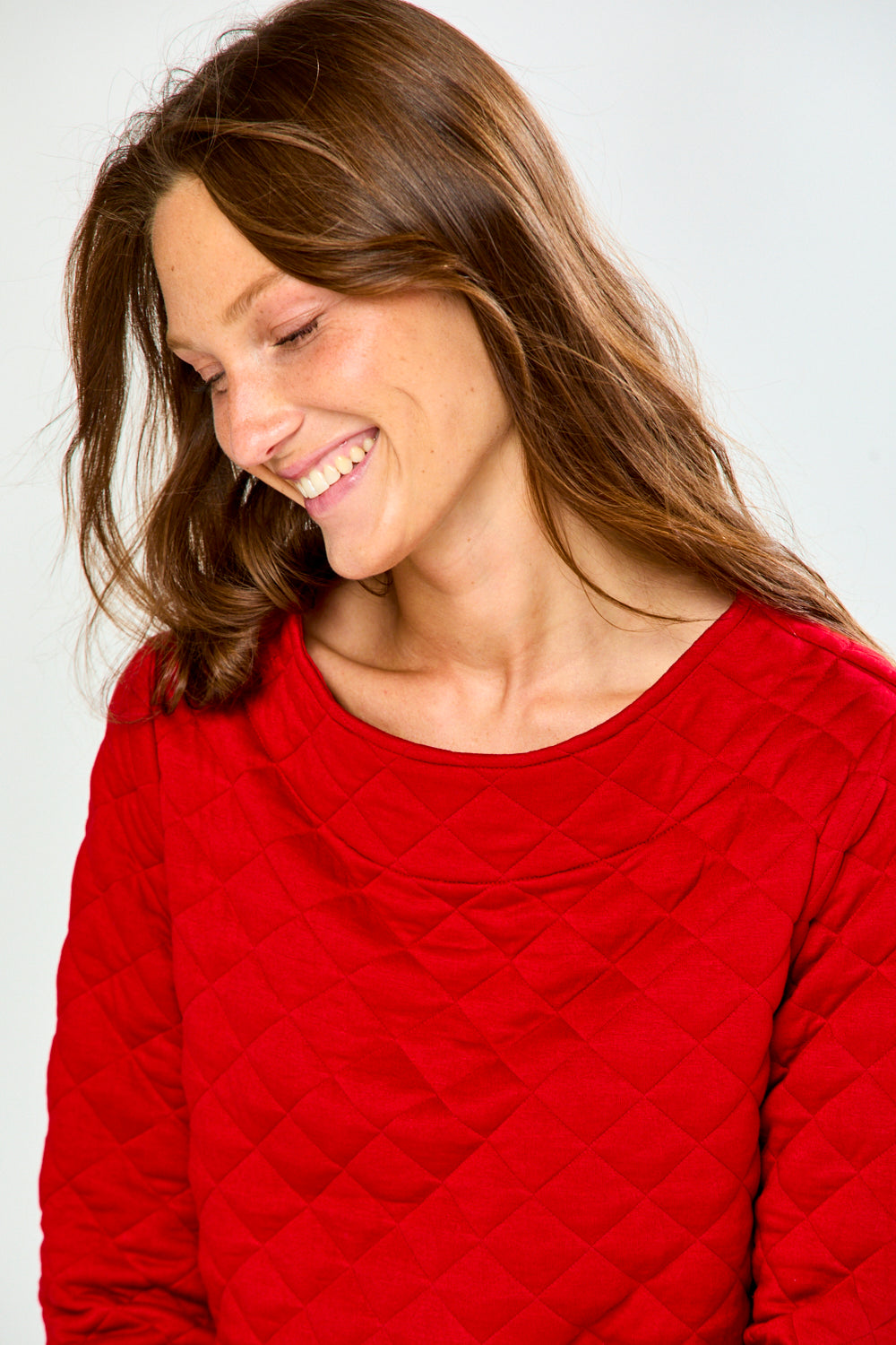 A woman modeling a red long-sleeve quilted knit top with a boat neckline, paired with green and red plaid pants, against a light gray studio background.