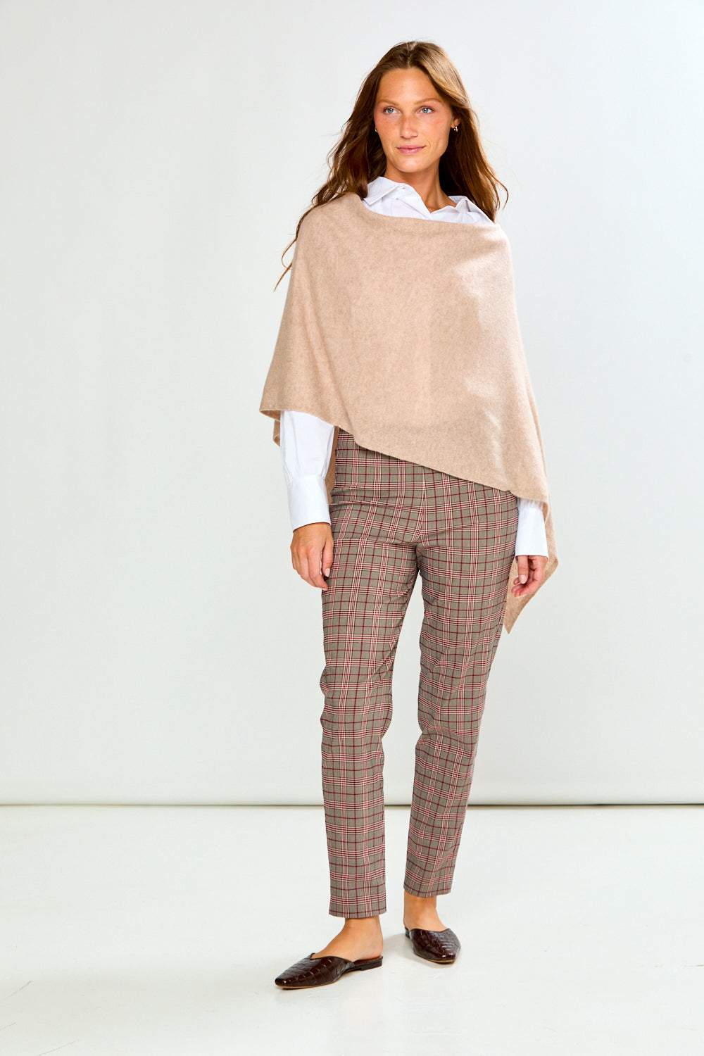 A woman modeling a light beige poncho draped over her shoulders, worn over a white collared shirt and paired with brown plaid trousers, against a plain light gray studio background.