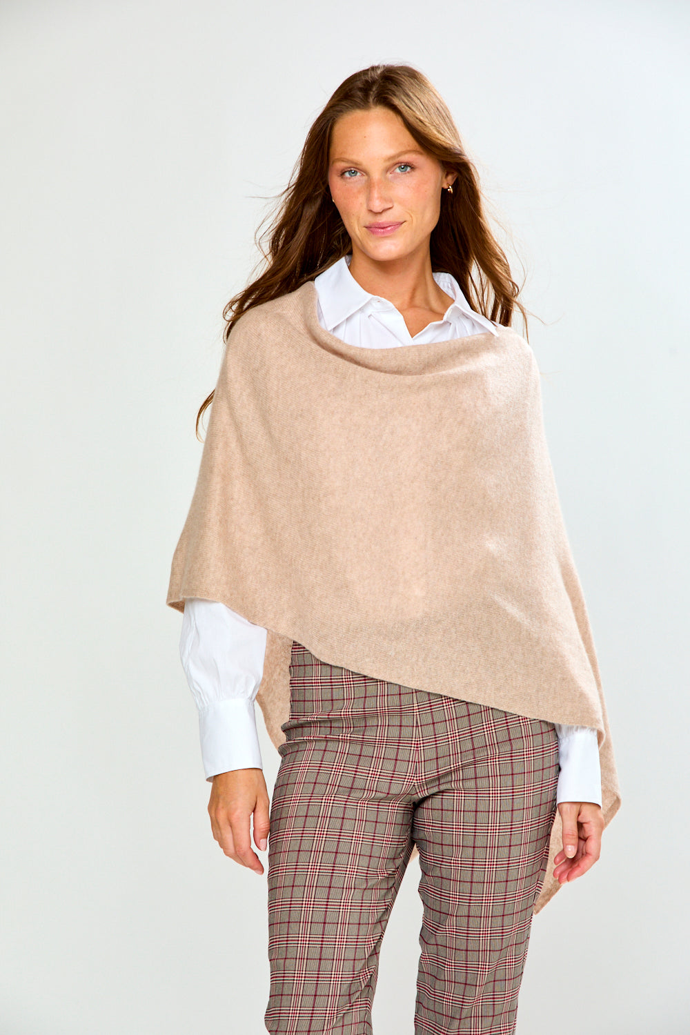 A woman modeling a light beige poncho draped over her shoulders, worn over a white collared shirt and paired with brown plaid trousers, against a plain light gray studio background.