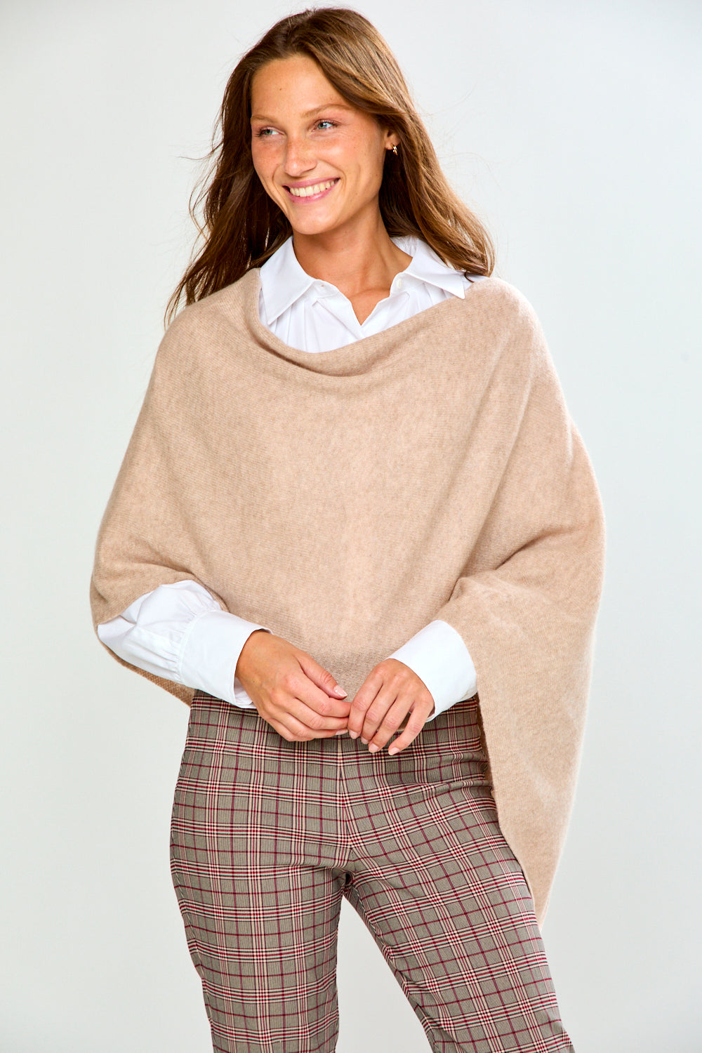 A woman modeling a light beige poncho draped over her shoulders, worn over a white collared shirt and paired with brown plaid trousers, against a plain light gray studio background.