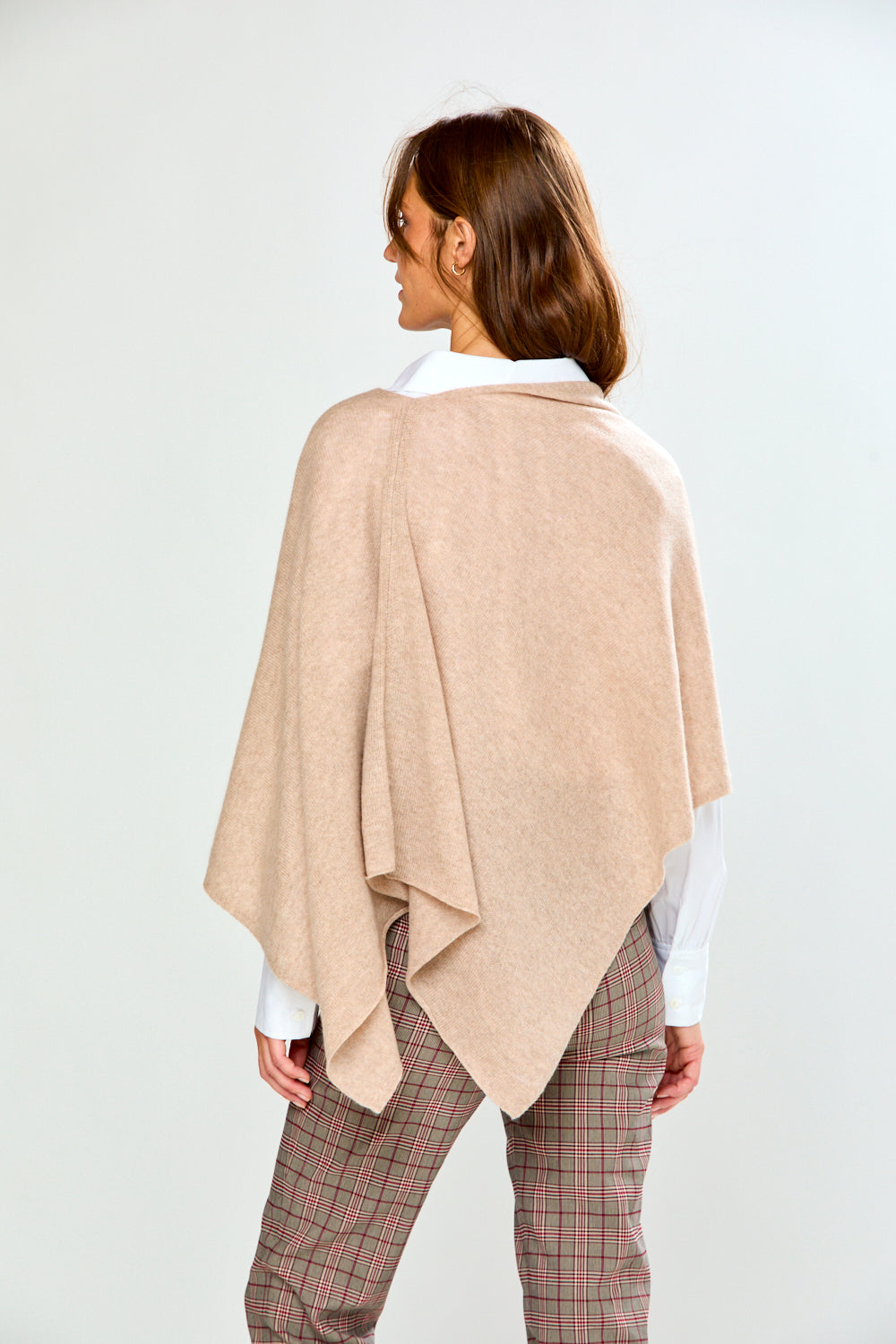 A woman modeling a light beige poncho draped over her shoulders, worn over a white collared shirt and paired with brown plaid trousers, against a plain light gray studio background.