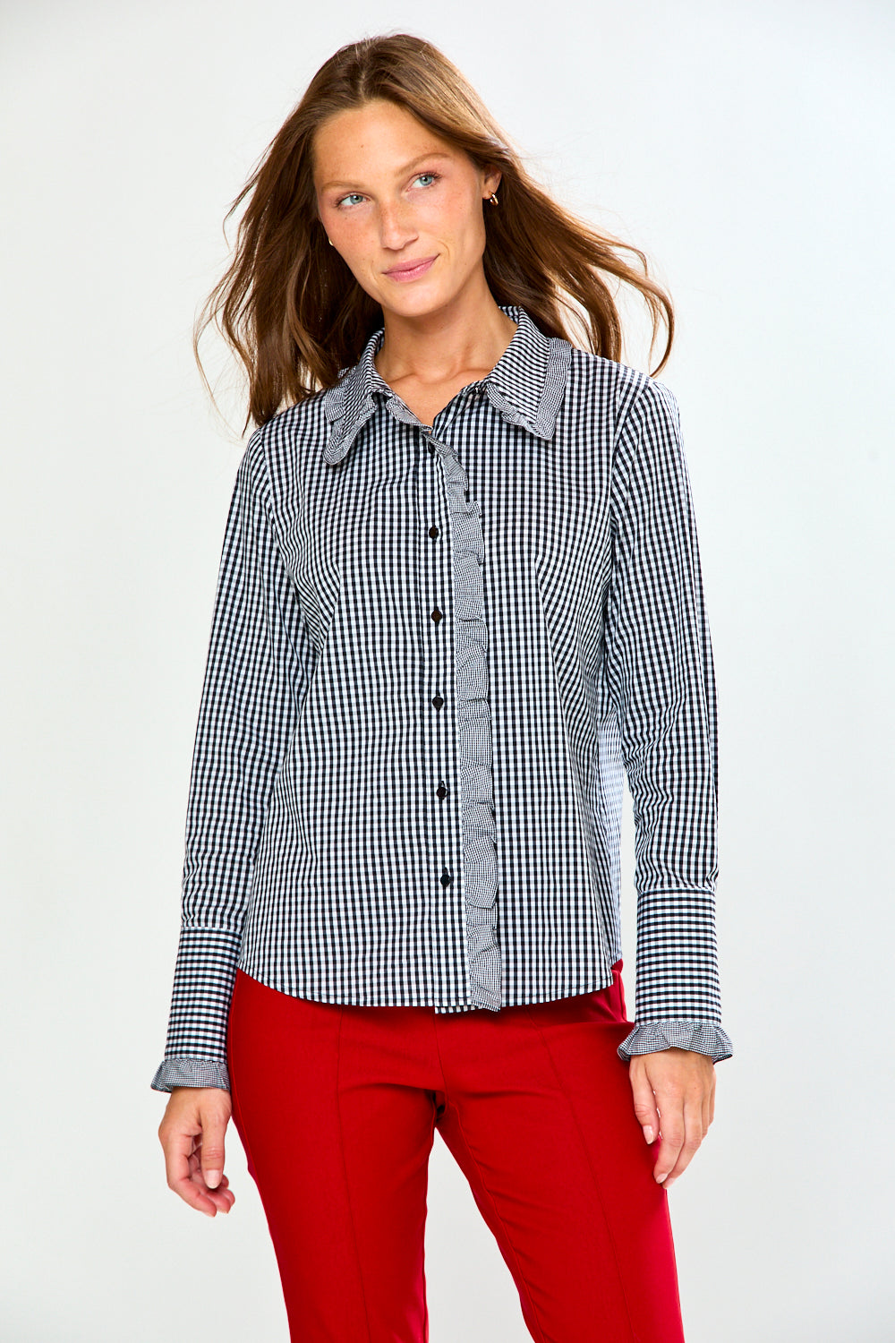 A woman modeling a long-sleeve button-up shirt with a black and white gingham pattern and a solid gray ruffled collar and placket, paired with red pants, against a plain white studio background.
