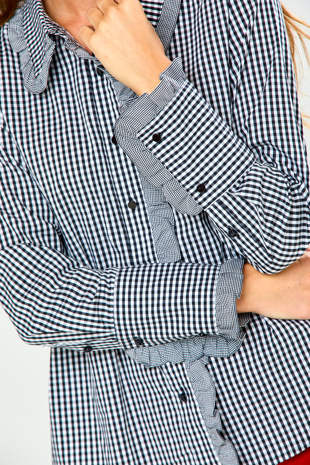 A close-up of a person wearing a black and white gingham long-sleeve shirt with ruffled trim along the collar, cuffs, and front opening. The image is cropped to show the torso and arms, with one hand near the neck and the other crossing the arm.