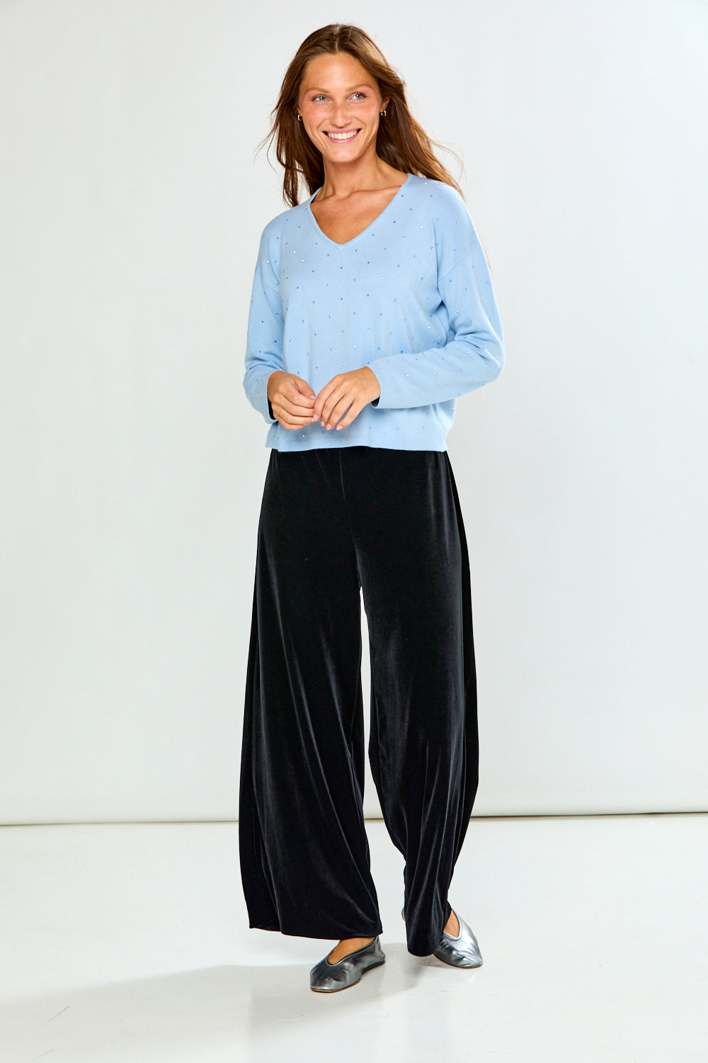 A woman modeling a light blue long-sleeve V-neck knit top with tiny crystal details, paired with black velvet pants, against a plain light gray studio background.