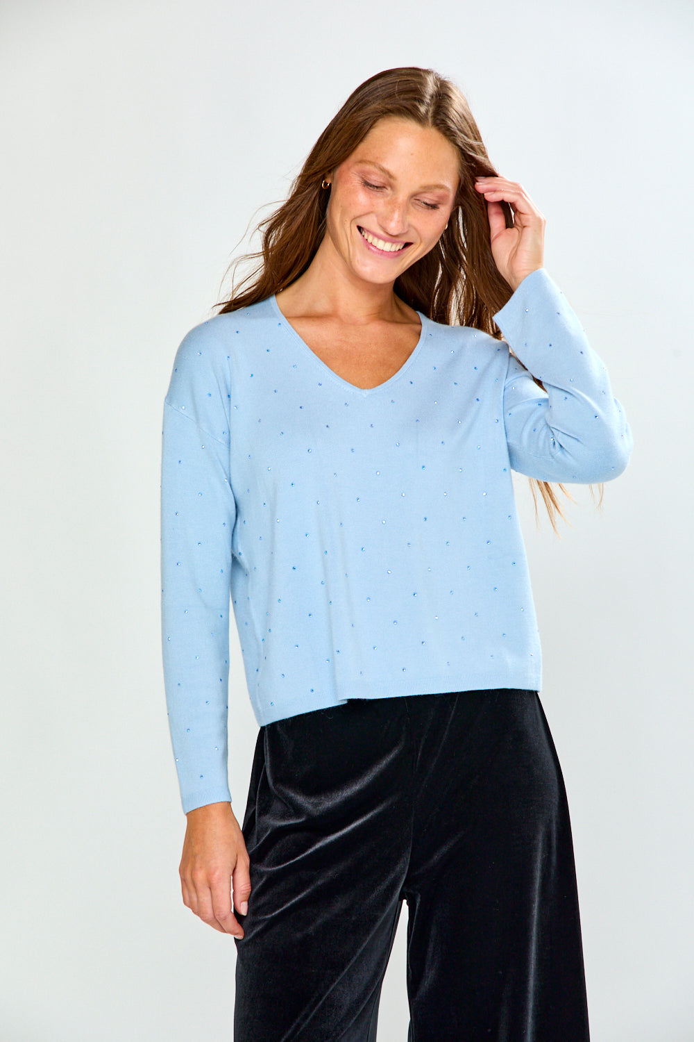 A woman modeling a light blue long-sleeve V-neck knit top with tiny crystal details, paired with black velvet pants, against a plain light gray studio background.