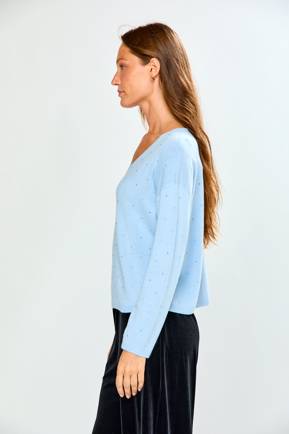 A woman modeling a light blue long-sleeve V-neck knit top with tiny crystal details, paired with black velvet pants, against a plain light gray studio background.