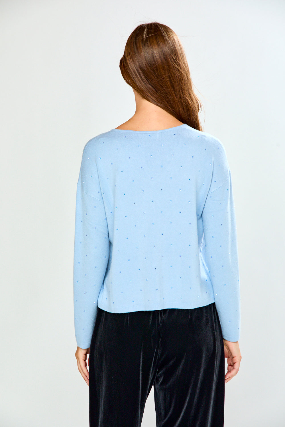 A woman modeling a light blue long-sleeve V-neck knit top with tiny crystal details, paired with black velvet pants, against a plain light gray studio background.