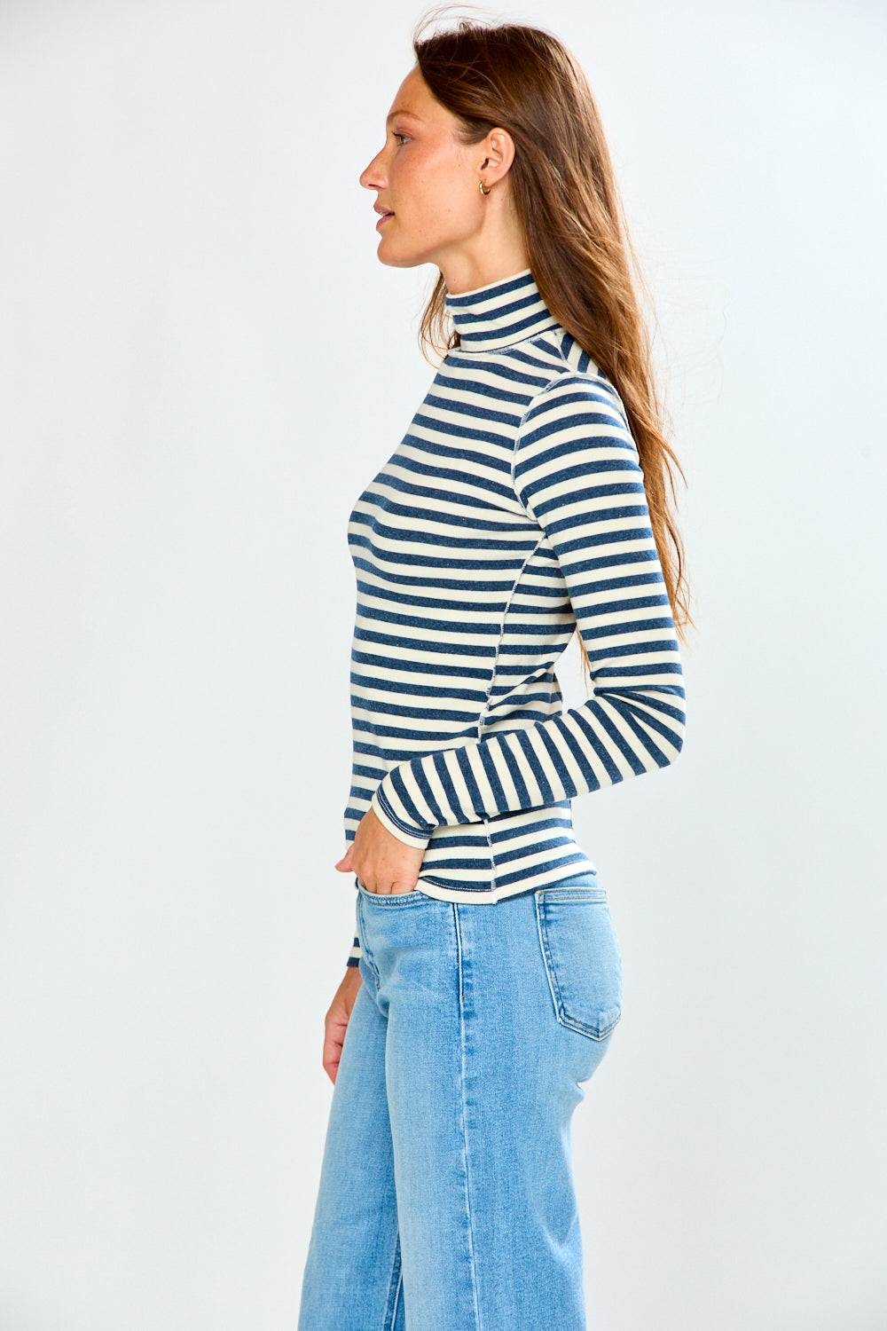 A woman wearing a navy and cream horizontal striped turtleneck sweater and light blue jeans, standing against a light gray studio background.