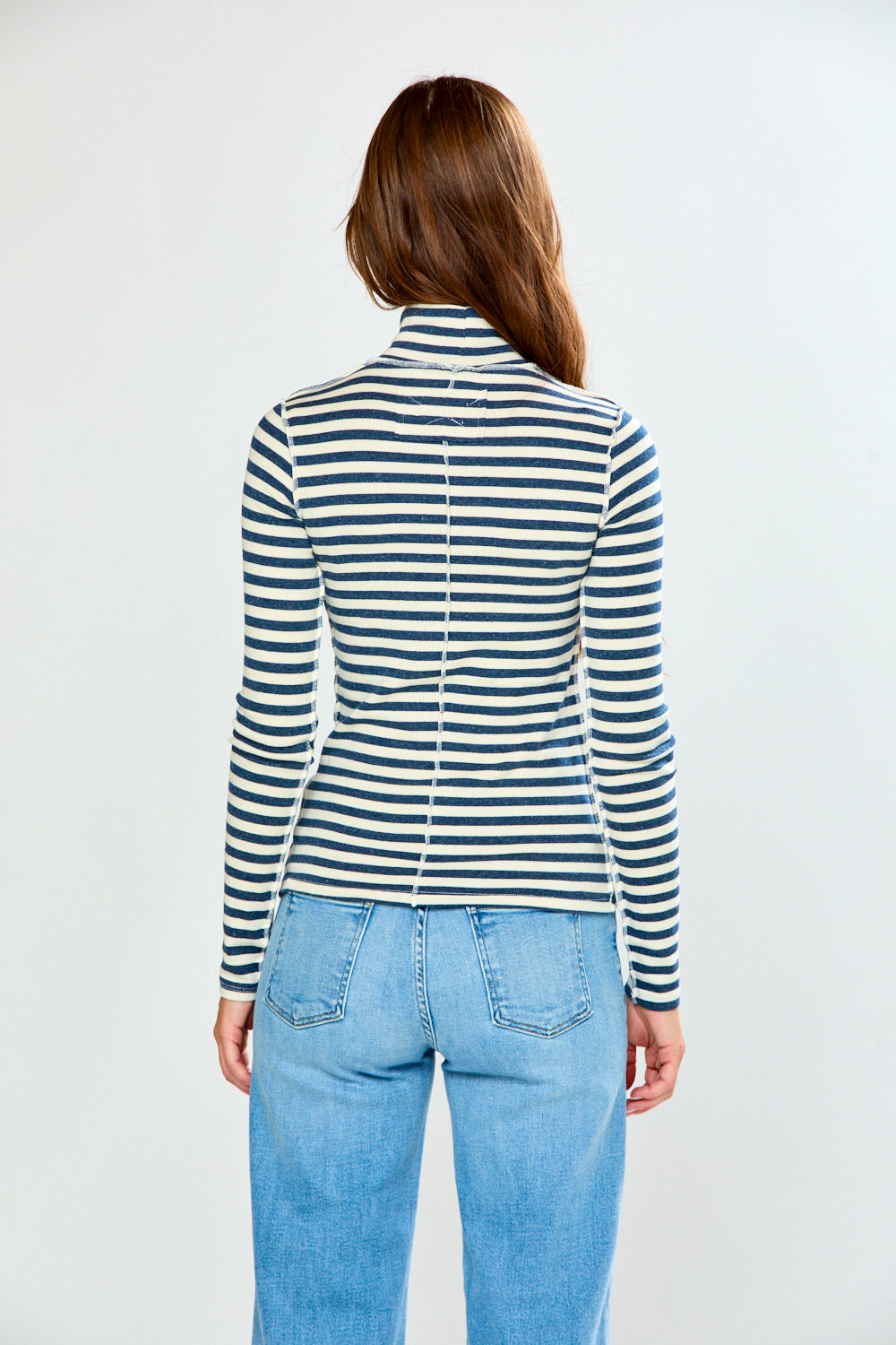 A woman wearing a navy and cream horizontal striped turtleneck sweater and light blue jeans, standing against a light gray studio background.