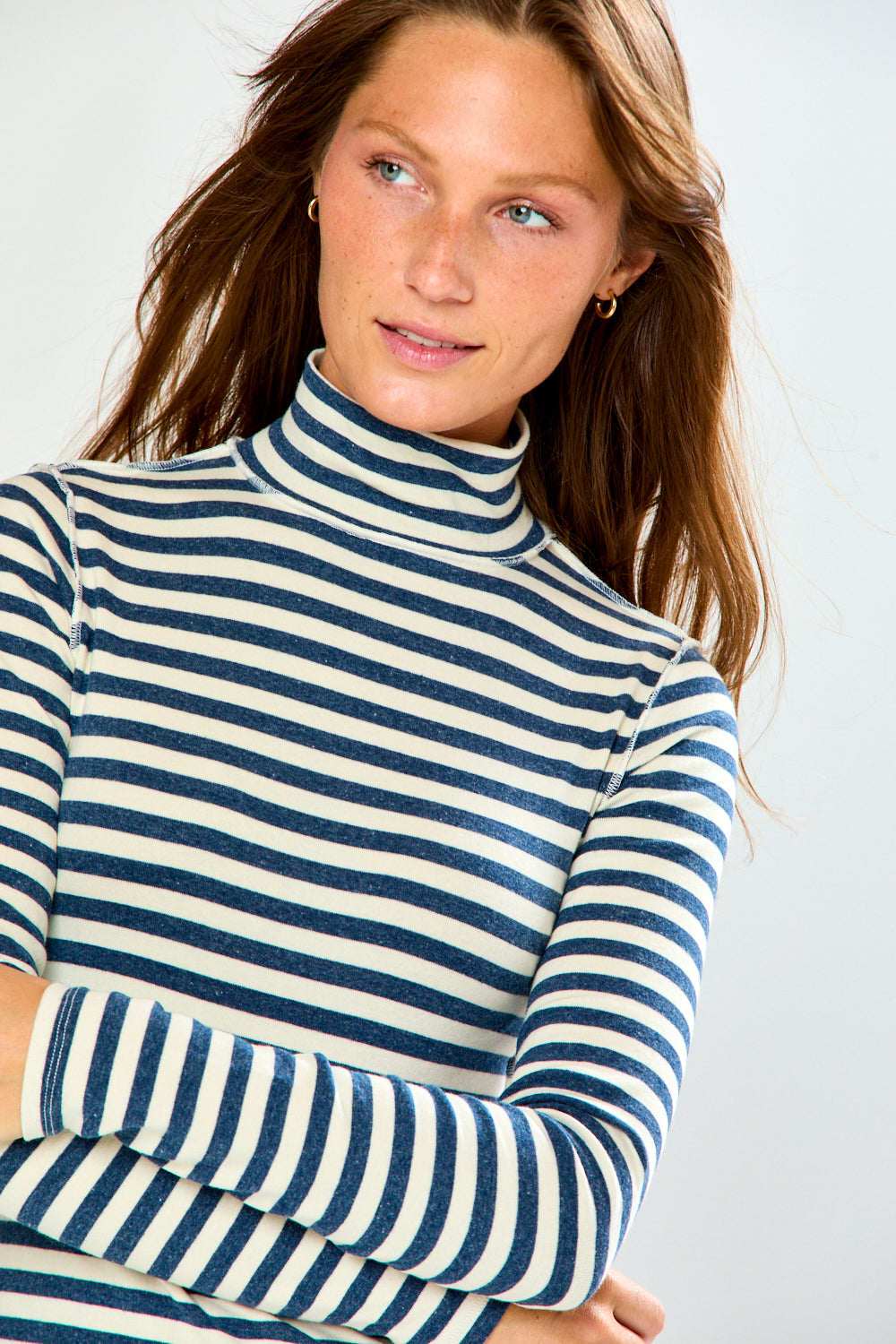 A woman wearing a navy and cream horizontal striped turtleneck sweater and light blue jeans, standing against a light gray studio background.