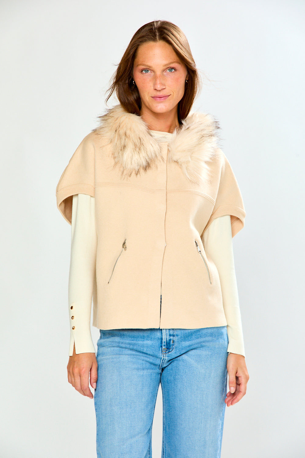 A woman modeling a beige sweater with a fluffy faux-fur collar and cream long sleeves, paired with light blue jeans, against a light gray studio background.