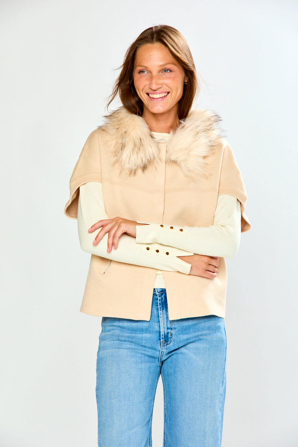 A woman modeling a beige sweater with a fluffy faux-fur collar and cream long sleeves, paired with light blue jeans, against a light gray studio background.
