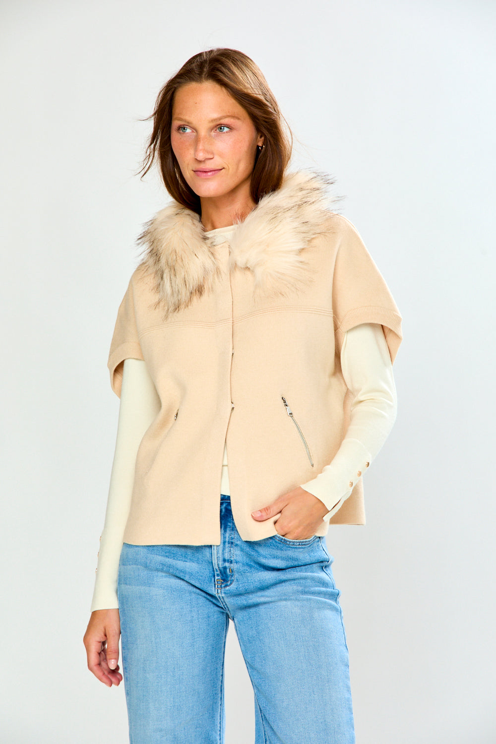 A woman modeling a beige sweater with a fluffy faux-fur collar and cream long sleeves, paired with light blue jeans, against a light gray studio background.