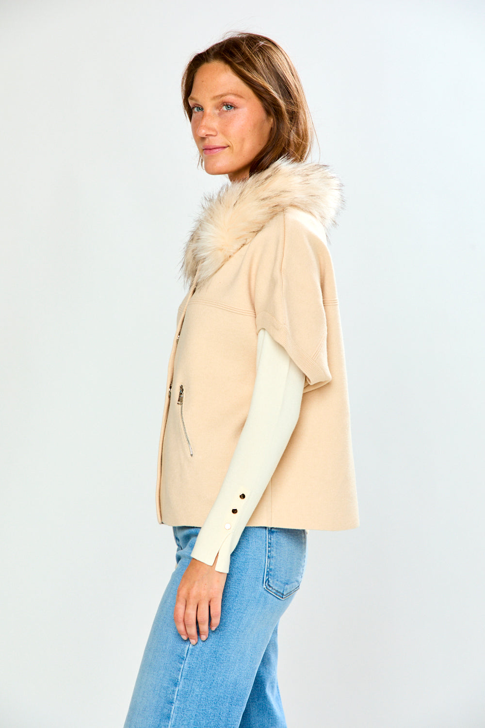 A woman modeling a beige sweater with a fluffy faux-fur collar and cream long sleeves, paired with light blue jeans, against a light gray studio background.
