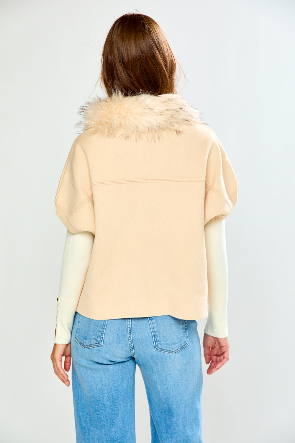 A woman modeling a beige sweater with a fluffy faux-fur collar and cream long sleeves, paired with light blue jeans, against a light gray studio background.