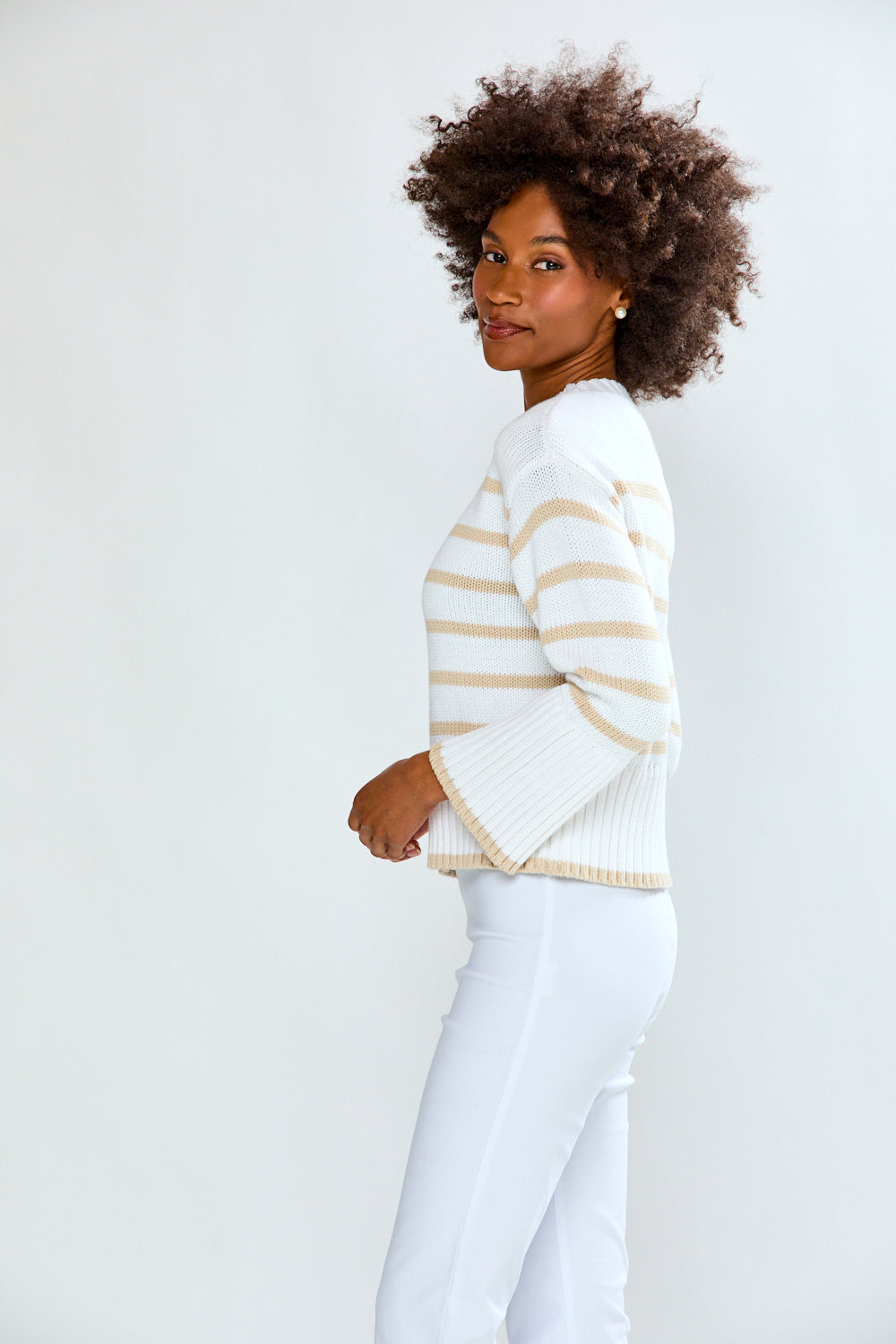 Woman wearing a white sweater with gold stripes and white pants on a plain background