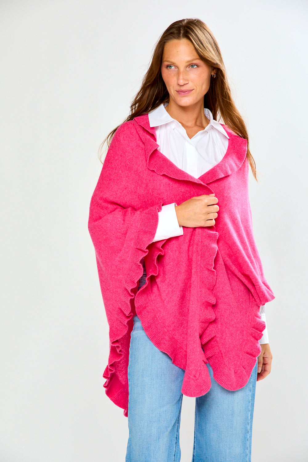A woman wearing a bright pink ruffled wrap over a white collared shirt and blue jeans, standing against a light gray studio background.