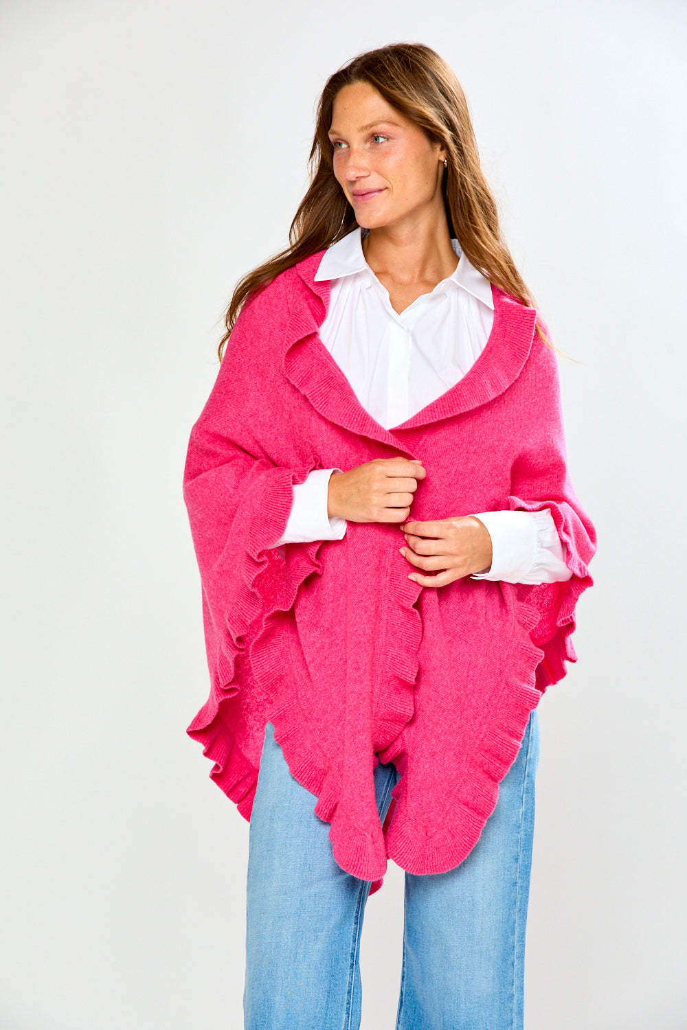 A woman wearing a bright pink ruffled wrap over a white collared shirt and blue jeans, standing against a light gray studio background.