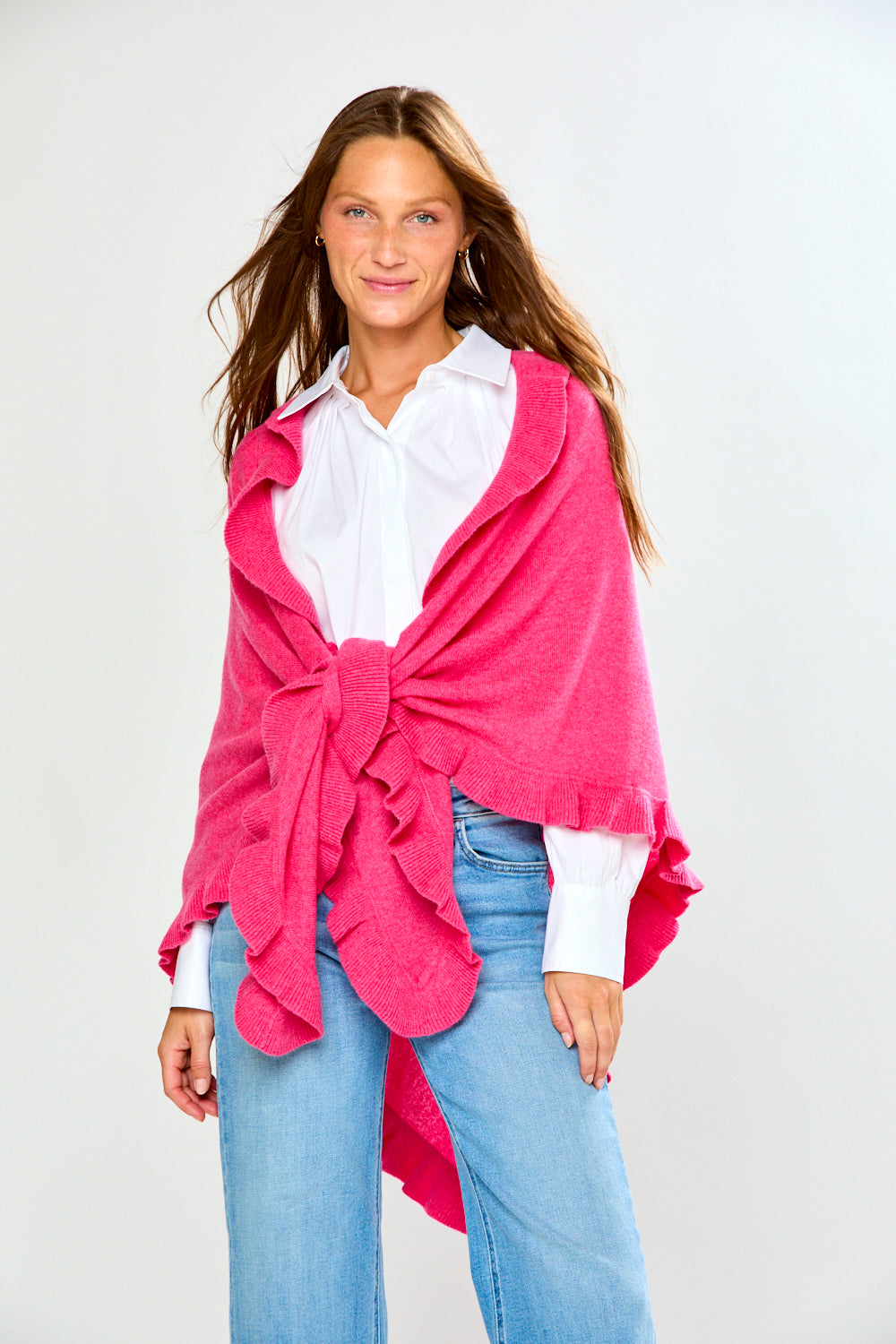 A woman wearing a bright pink ruffled wrap over a white collared shirt and blue jeans, standing against a light gray studio background.