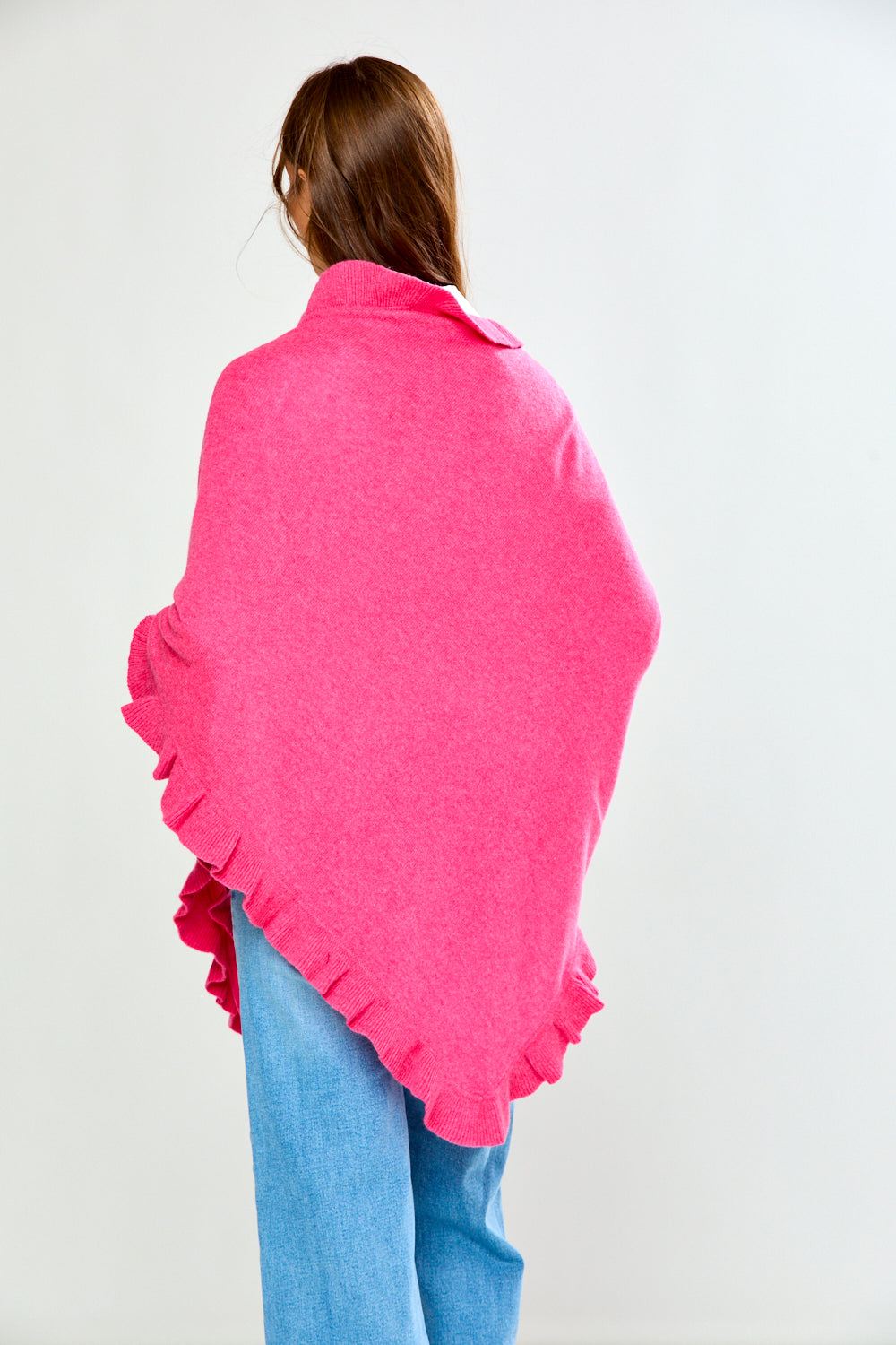 A woman wearing a bright pink ruffled wrap over a white collared shirt and blue jeans, standing against a light gray studio background.