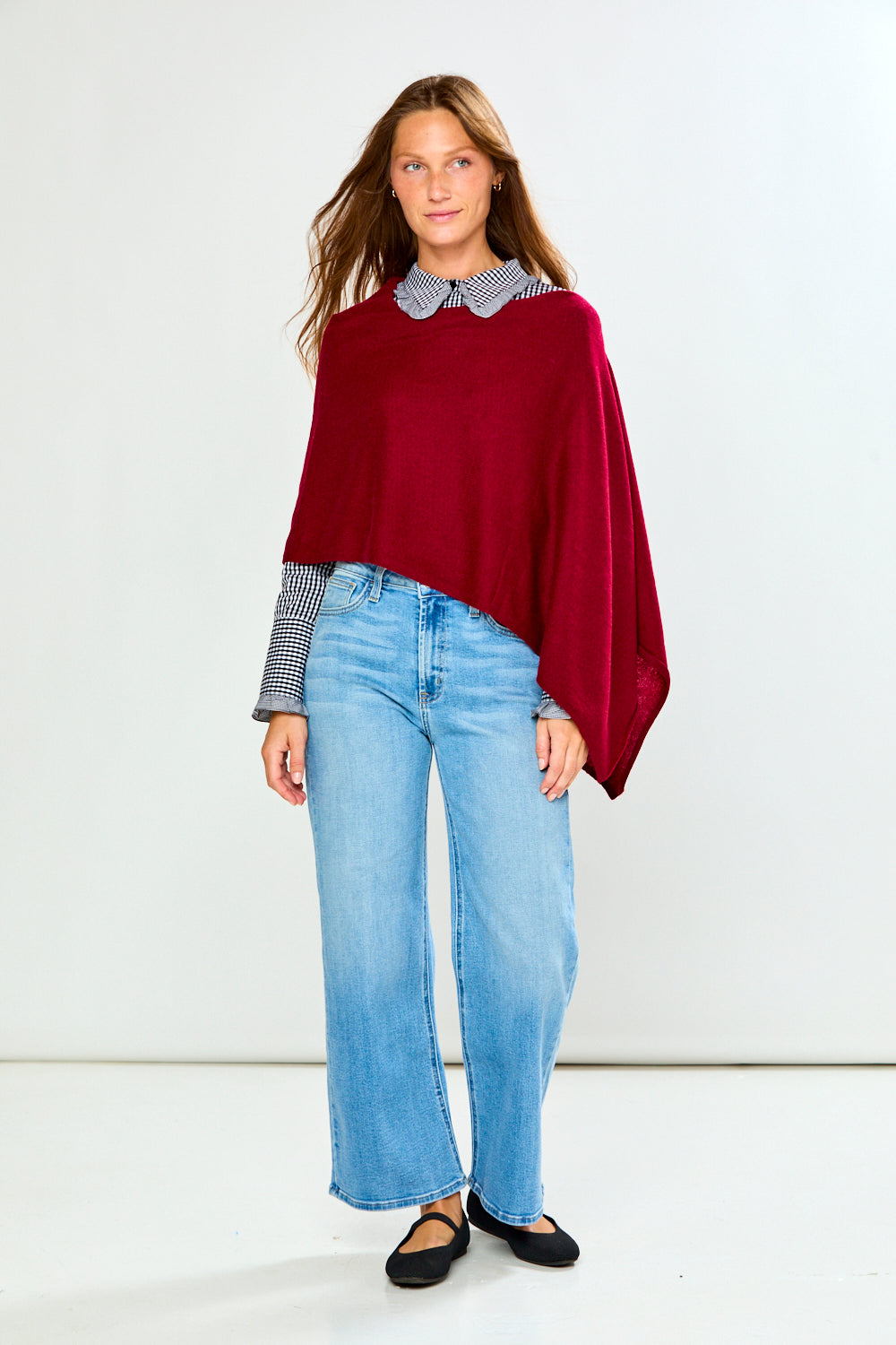 A woman modeling a burgundy knit poncho layered over a black and white gingham long-sleeve shirt, paired with light blue wide-leg jeans and black flats, against a plain white studio background.