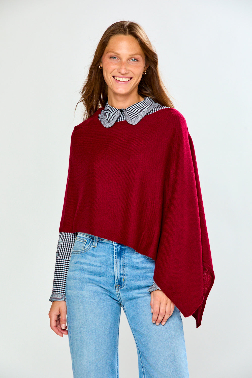 A woman modeling a deep burgundy knit poncho layered over a black and white gingham long-sleeve shirt with a pointed collar, paired with light blue jeans, against a light gray studio background.