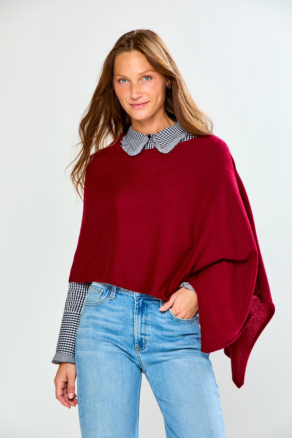 A woman modeling a deep burgundy knit poncho sweater with a black and white checkered Peter Pan collar, worn over a matching checkered shirt and paired with light blue jeans, against a light gray studio background.