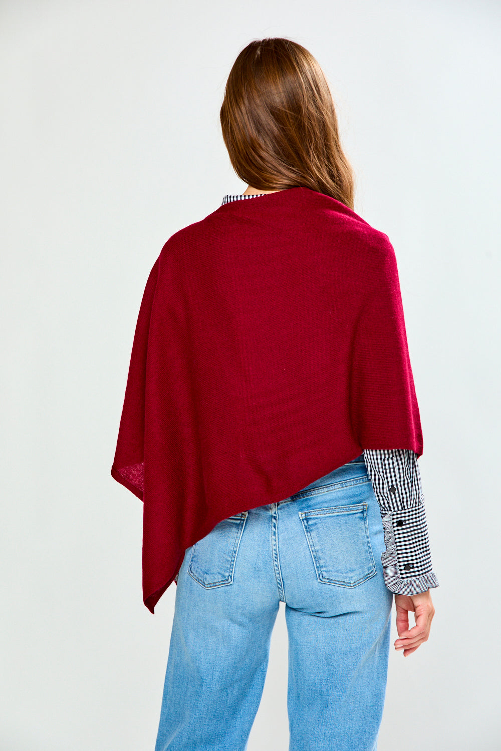 A woman shown from behind wearing a deep burgundy poncho over a black and white checkered shirt and light blue jeans, set against a plain light gray studio background.