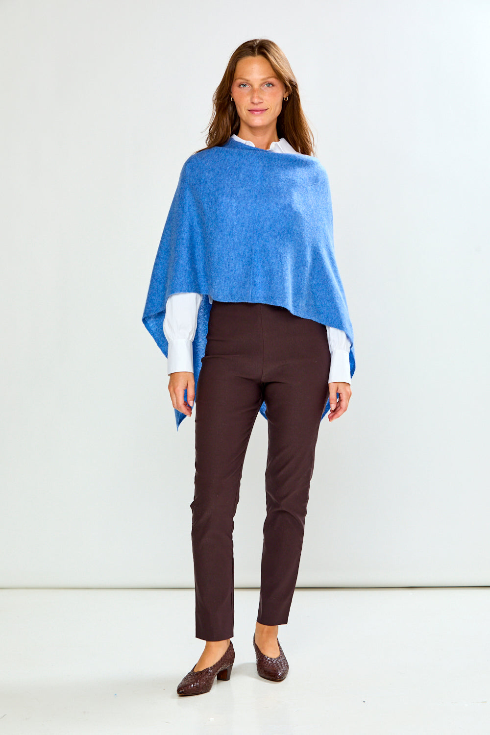 A woman modeling a bright blue poncho over a white collared shirt and dark brown trousers, standing against a light gray studio background.