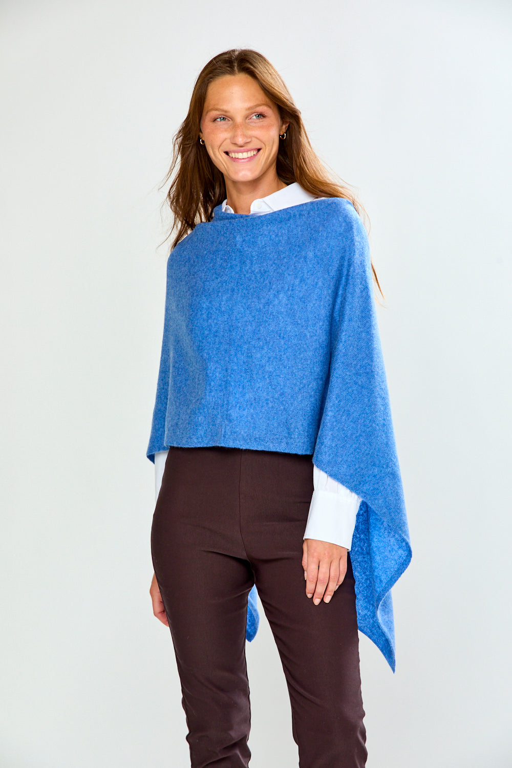 A woman modeling a bright blue poncho over a white collared shirt and dark brown trousers, standing against a light gray studio background.