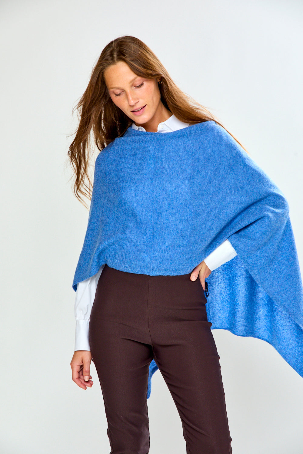 A woman modeling a bright blue poncho over a white collared shirt and dark brown trousers, standing against a light gray studio background.