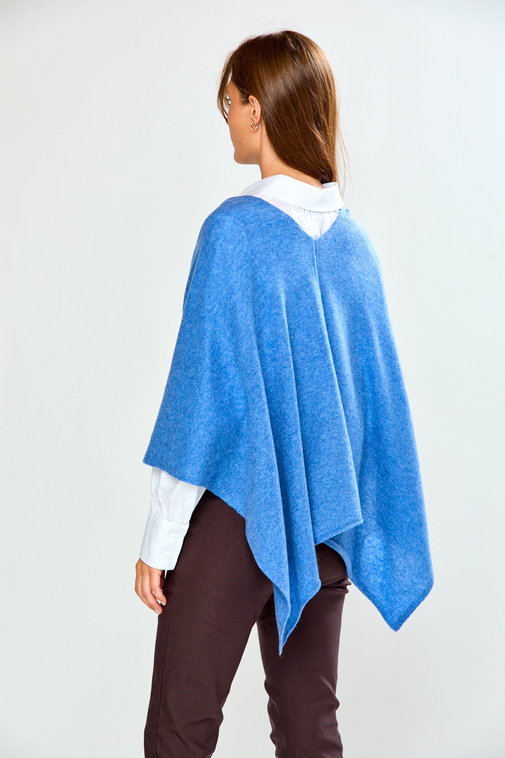 A woman modeling a bright blue poncho over a white collared shirt and dark brown trousers, standing against a light gray studio background.