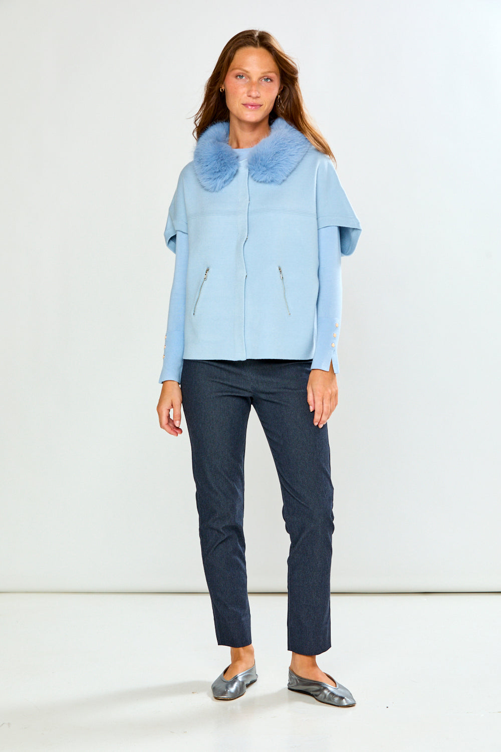 A woman modeling a light blue cropped sweater with a fluffy light blue faux-fur collar, paired with dark gray trousers, against a plain light gray studio background.