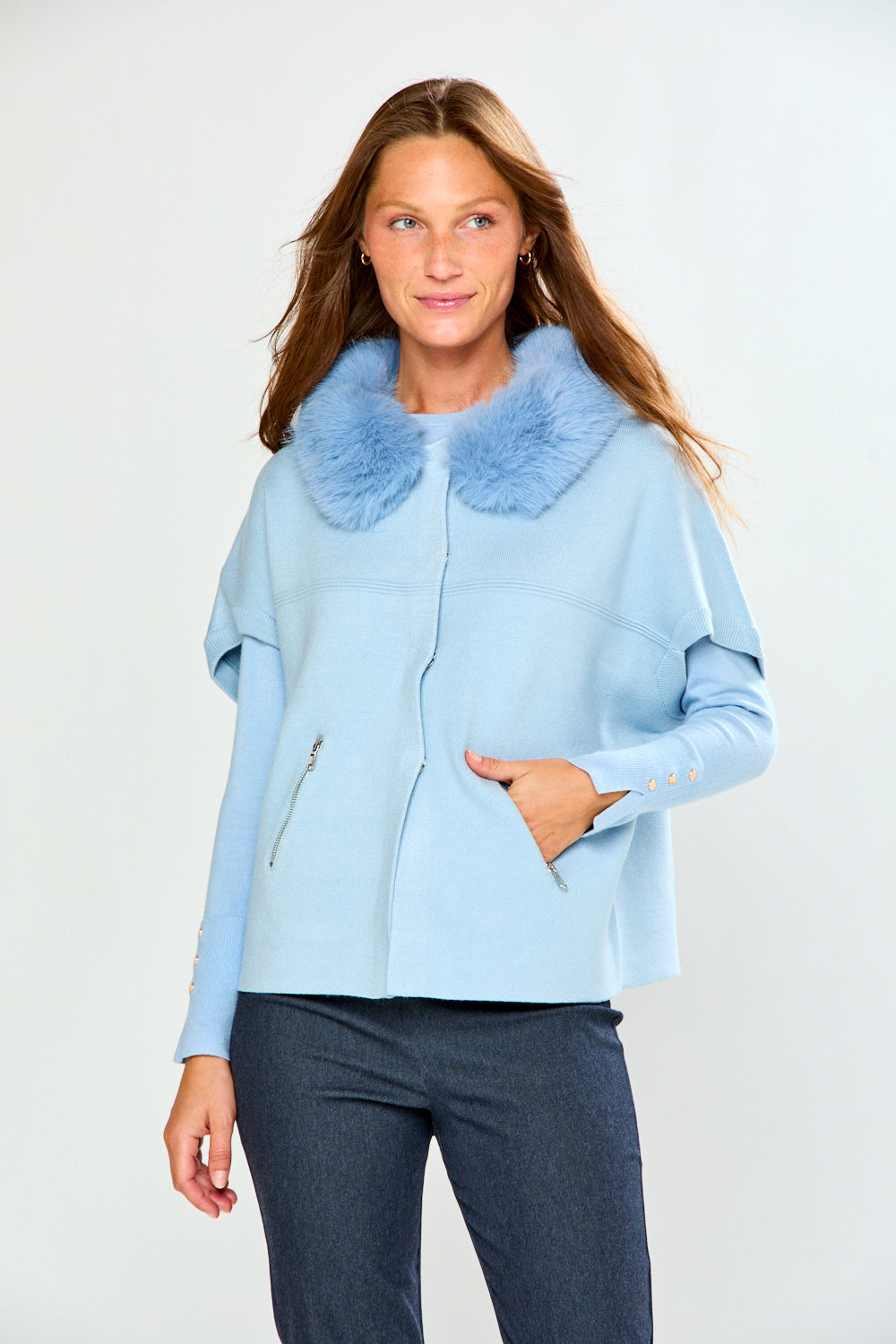 A woman modeling a light blue cropped sweater with a fluffy light blue faux-fur collar, paired with dark gray trousers, against a plain light gray studio background.