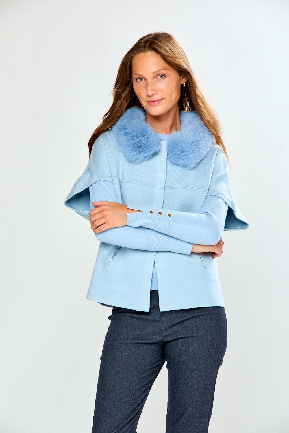 A woman modeling a light blue cropped sweater with a fluffy light blue faux-fur collar, paired with dark gray trousers, against a plain light gray studio background.