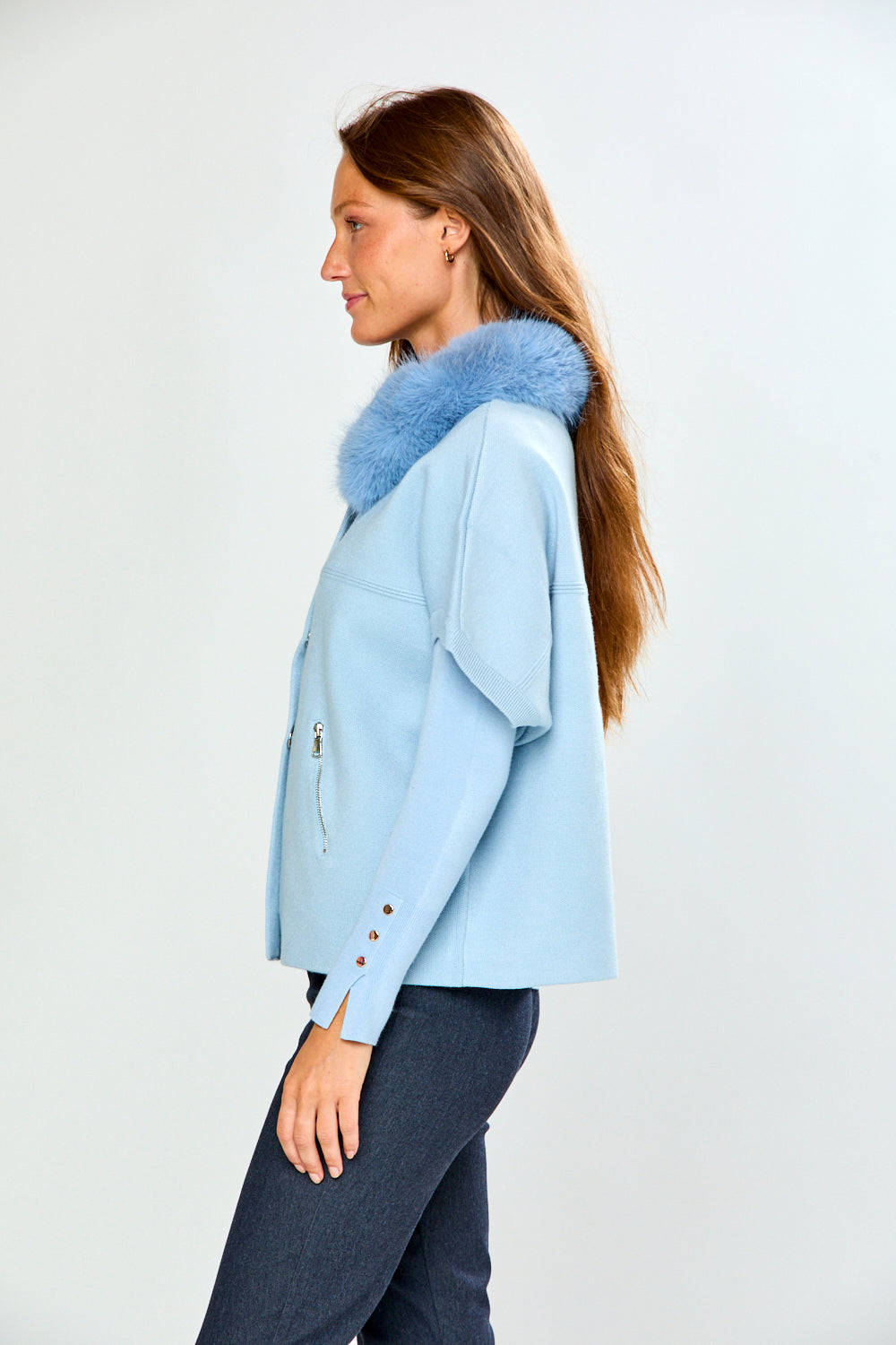 A woman modeling a light blue cropped sweater with a fluffy light blue faux-fur collar, paired with dark gray trousers, against a plain light gray studio background.