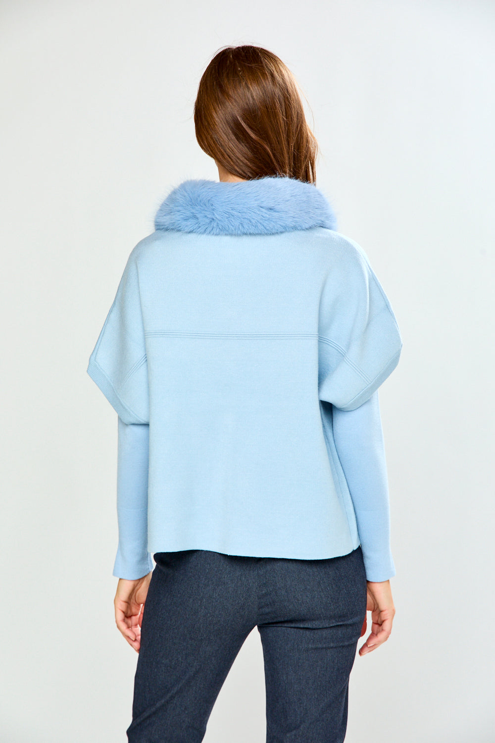 A woman modeling a light blue cropped sweater with a fluffy light blue faux-fur collar, paired with dark gray trousers, against a plain light gray studio background.