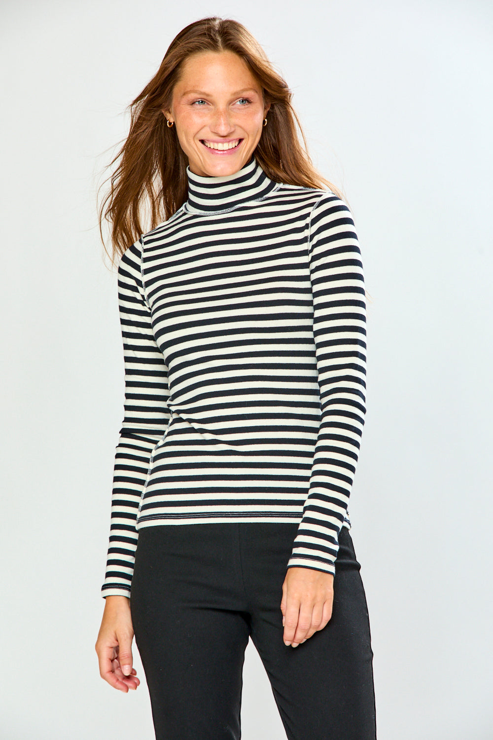A woman wearing a black and white horizontally striped long-sleeve turtleneck sweater, paired with black pants, standing against a light gray studio background.