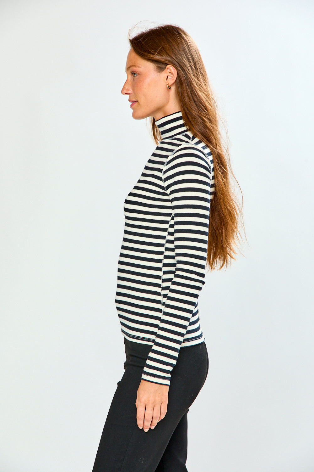 A woman in profile wearing a black and white horizontally striped mock neck turtleneck sweater and black pants, standing against a light gray studio background.
