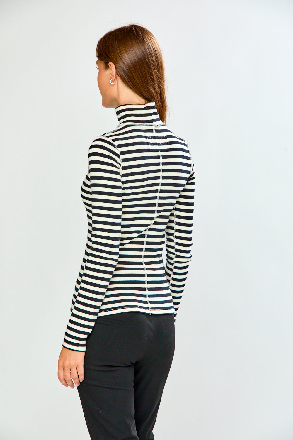 A woman shown from the back wearing a black and white horizontally striped long-sleeve turtleneck top and black pants, against a plain light gray studio background.