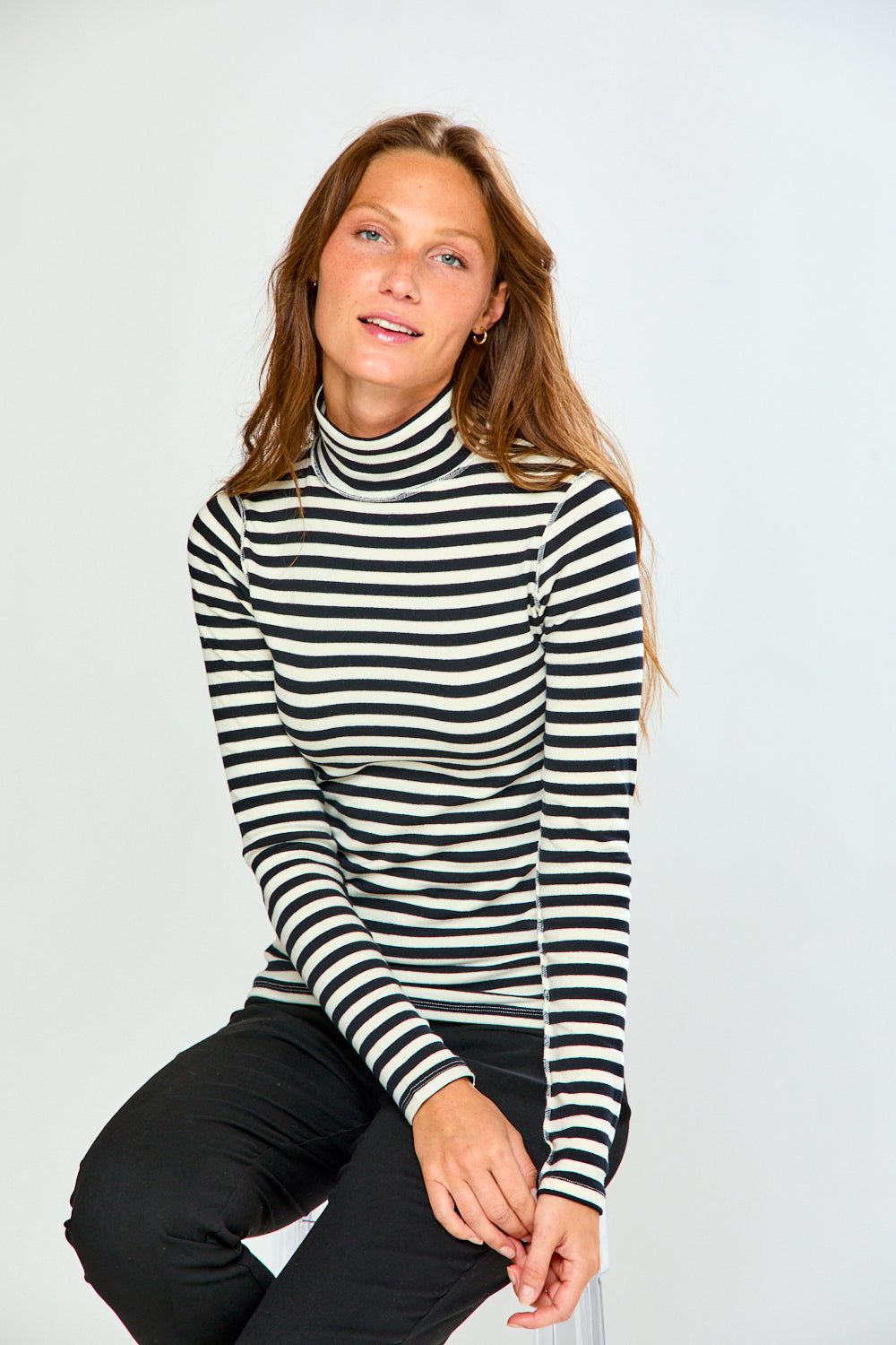 A woman wearing a black and white horizontally striped long-sleeve turtleneck sweater, paired with black pants, posing against a light gray studio background.