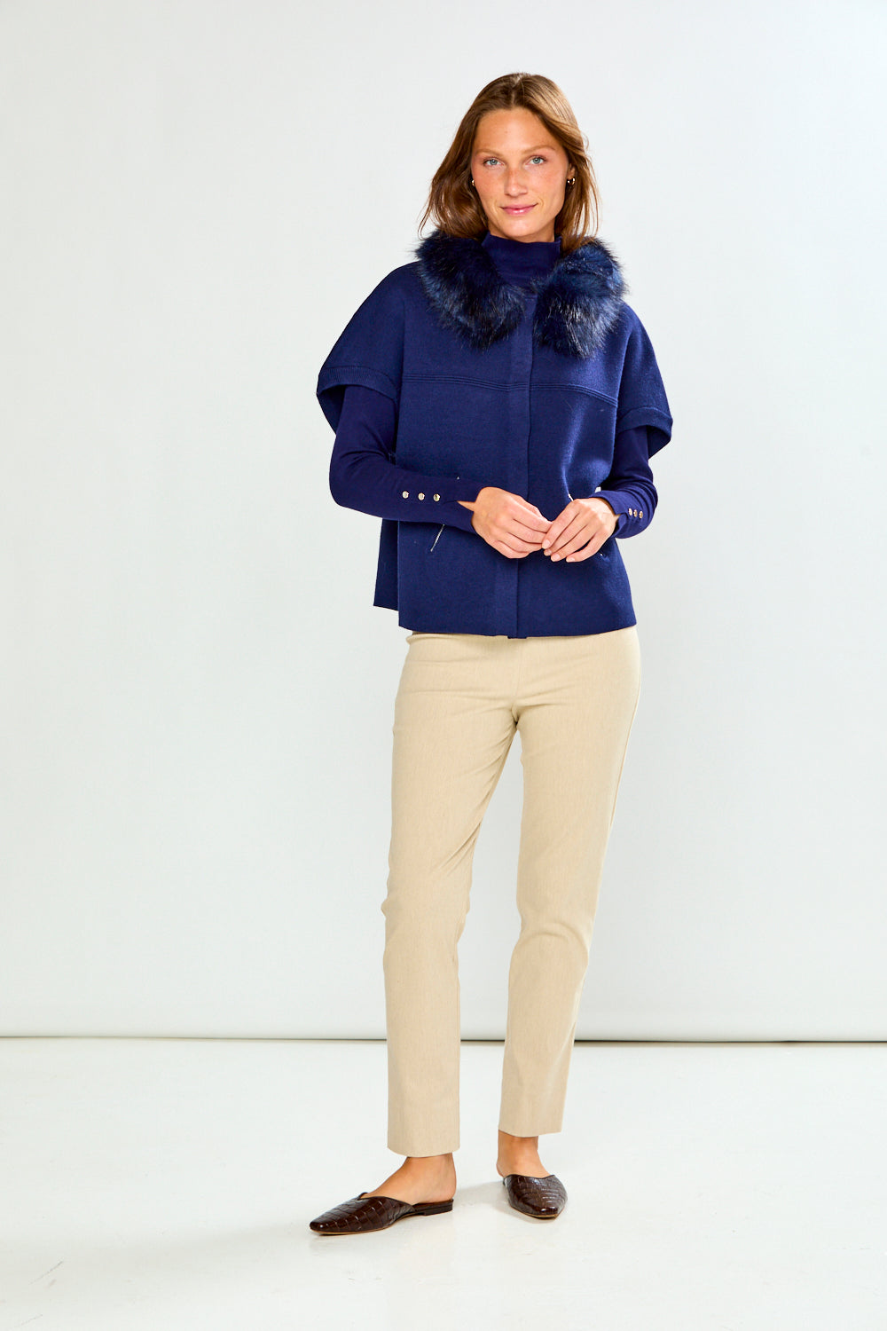 A woman modeling a navy blue cropped sweater with a dark fur-trimmed collar and a diagonal zipper pocket, paired with beige pants, against a light gray studio background.