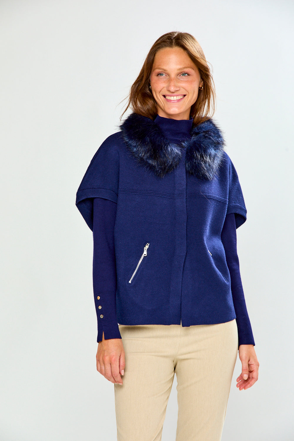 A woman modeling a navy blue cropped sweater with a dark fur-trimmed collar and a diagonal zipper pocket, paired with beige pants, against a light gray studio background.