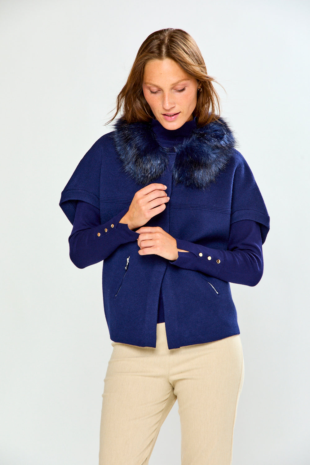 A woman wearing a navy blue jacket with a dark blue fur collar and beige pants, standing against a light gray studio background.