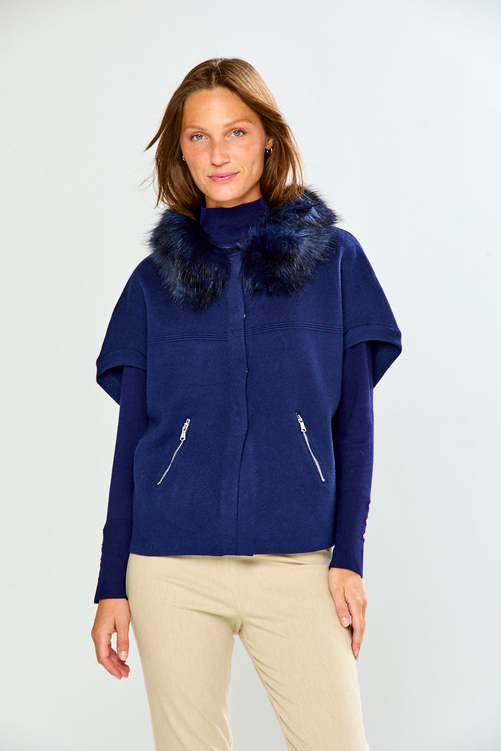 A woman modeling a navy blue jacket with a dark blue fur-trimmed collar and silver zipper details, paired with beige pants, against a light gray studio background.