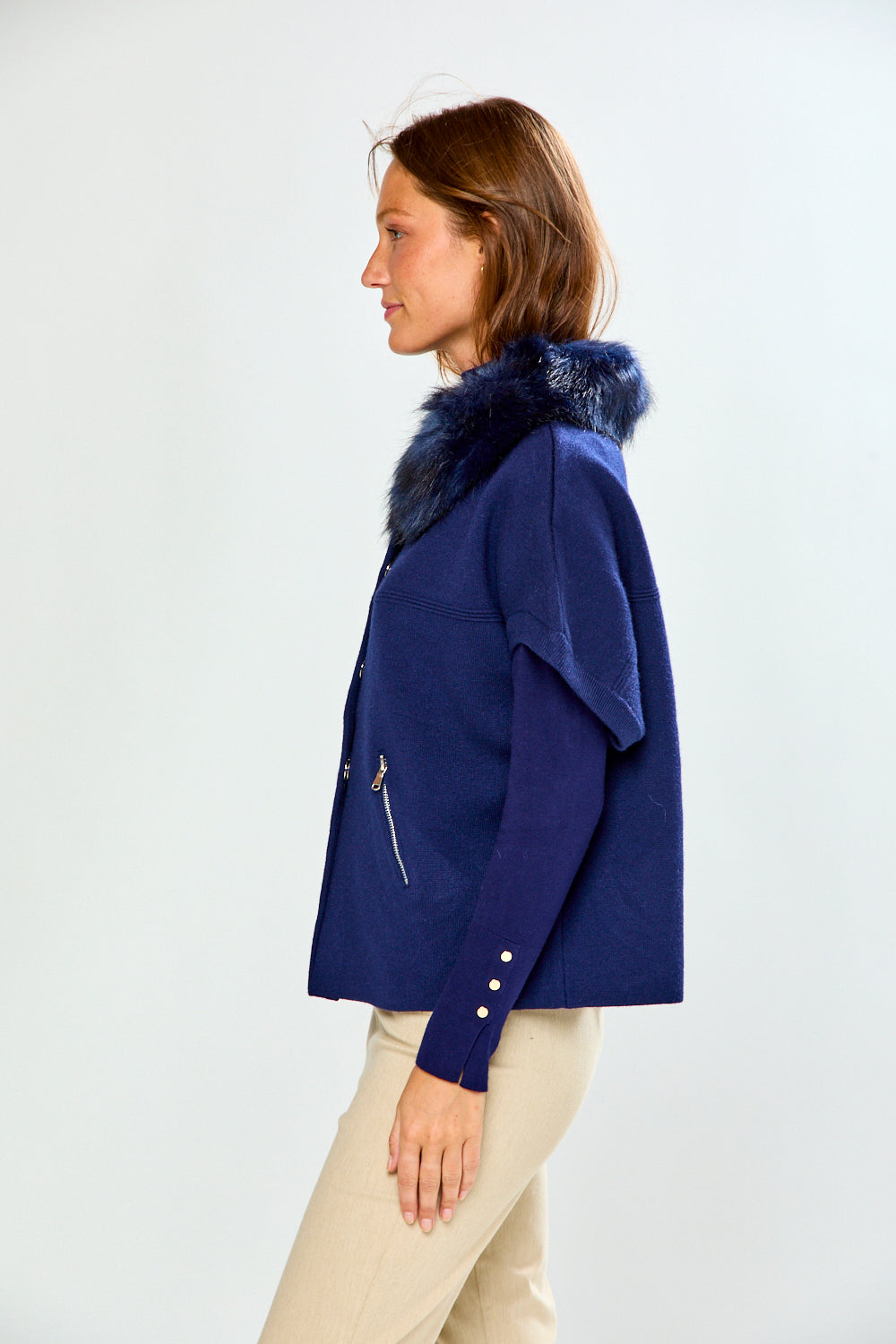 A woman modeling a navy blue cropped sweater with a dark fur-trimmed collar and a diagonal zipper pocket, paired with beige pants, against a light gray studio background.
