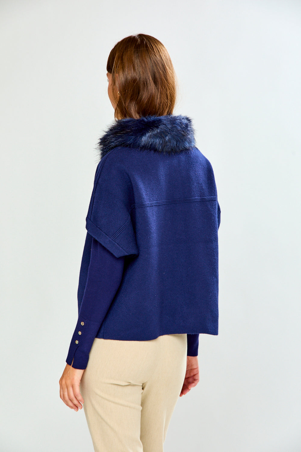 A woman shown from the back wearing a navy blue cropped jacket with a dark blue faux-fur collar, paired with beige trousers, against a plain light gray background.