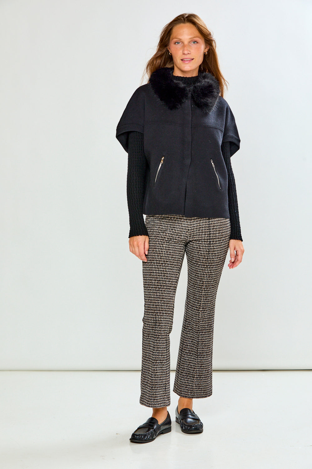 A woman modeling a black sleeveless sweater with a fluffy black faux-fur collar, worn over a black ribbed long-sleeve top and paired with black-and-white houndstooth pants, against a light gray studio background.