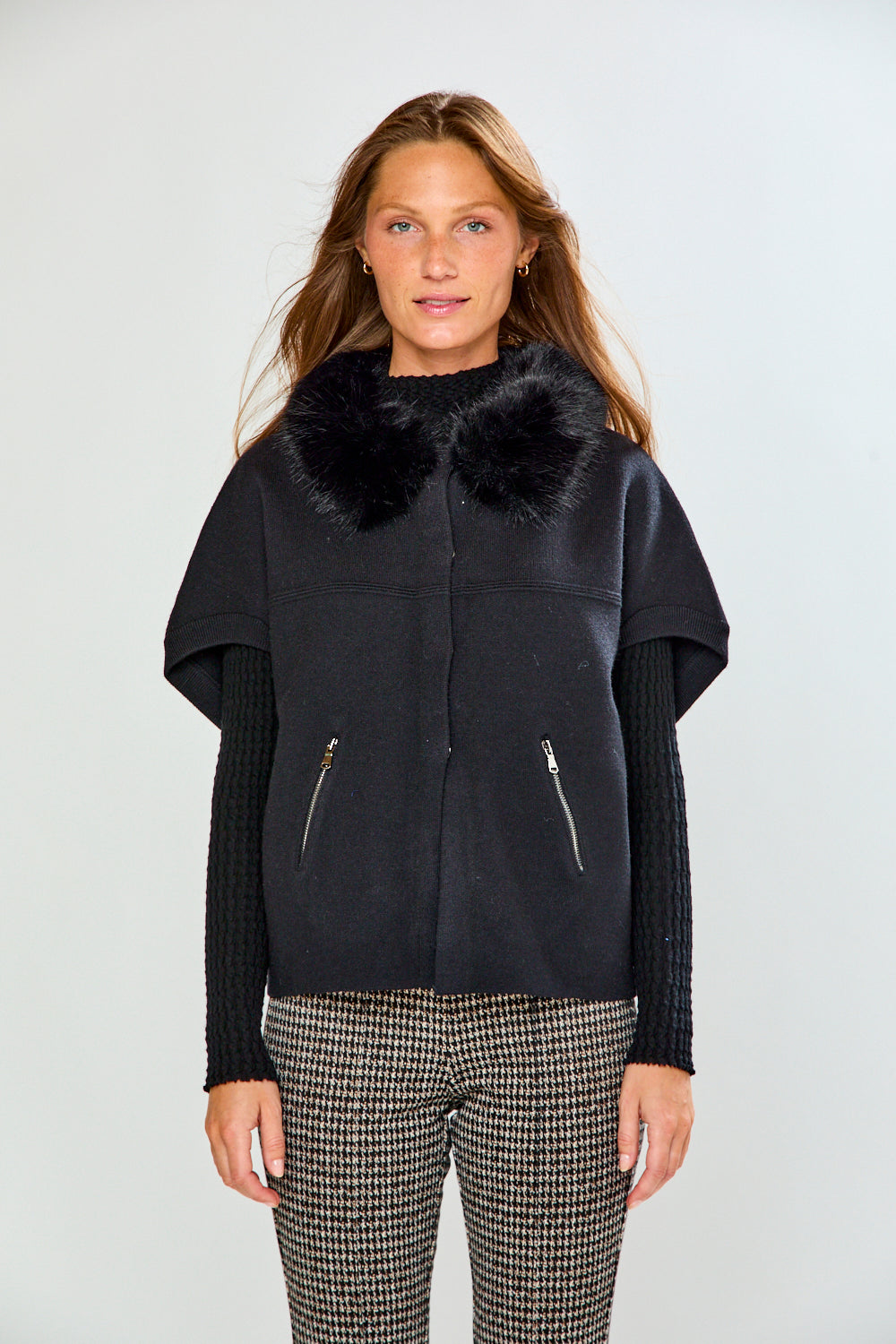 A woman modeling a black sleeveless sweater with a fluffy black faux-fur collar, worn over a black ribbed long-sleeve top and paired with black-and-white houndstooth pants, against a light gray studio background.